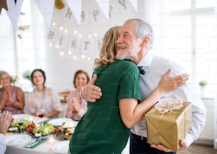 Unrecognizable young woman giving a gift to her father or grandfather on indoor party, hugging.
