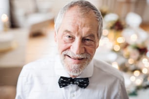 A close-up portrait of a senior man with gray beard and mustache standing indoors in a room set for a party.