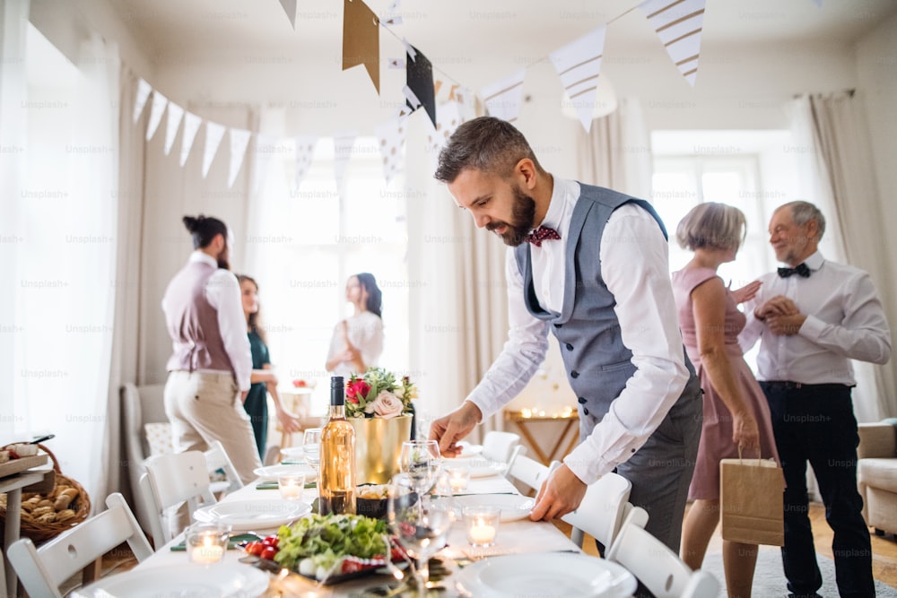Un uomo maturo con fiocco e gilet che apparecchia un tavolo per una festa al coperto, ospiti sullo sfondo.