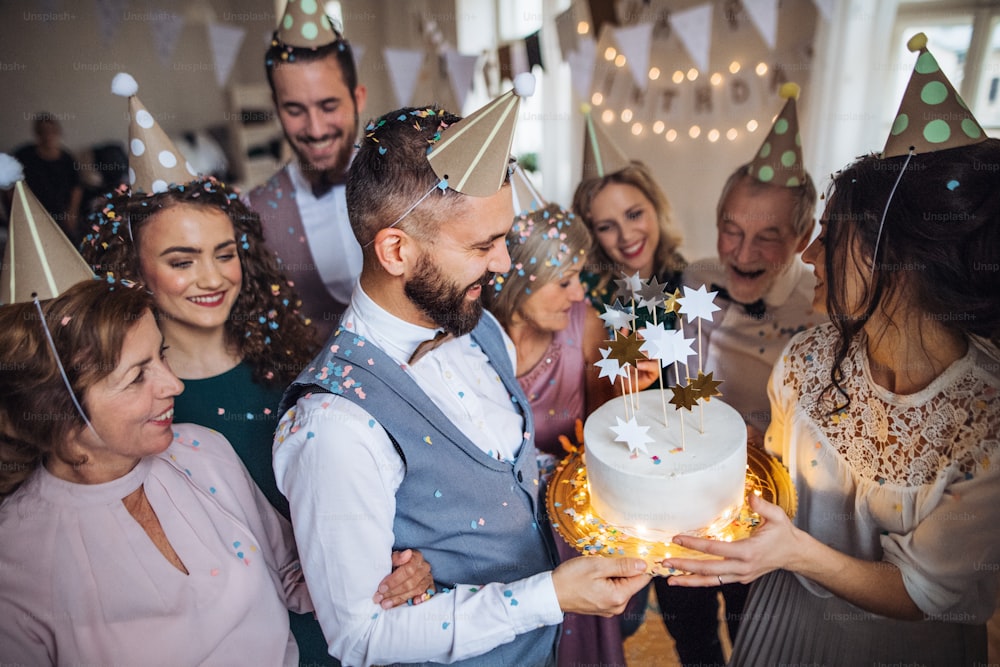 Un retrato de una familia multigeneracional con un pastel y sombreros de fiesta en una fiesta de cumpleaños en el interior.