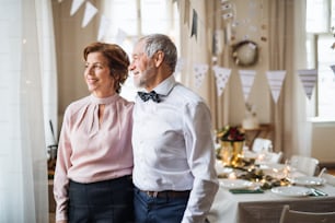 A portrait of a senior couple standing indoors in a room set for a party. Copy space.