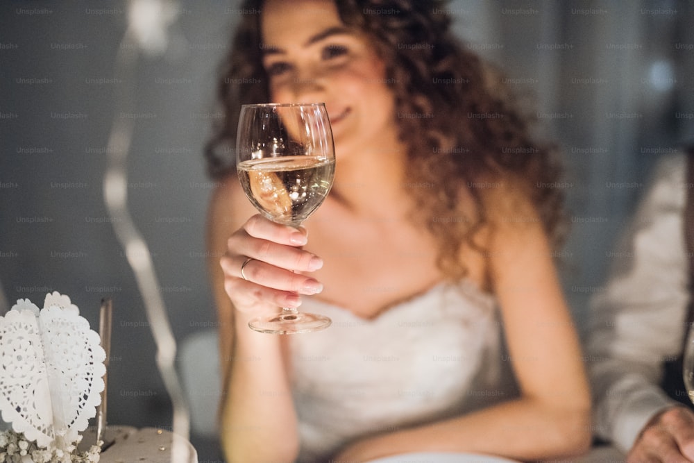 Um close-up de uma jovem noiva sentada em uma mesa em um casamento, segurando um copo de vinho branco.