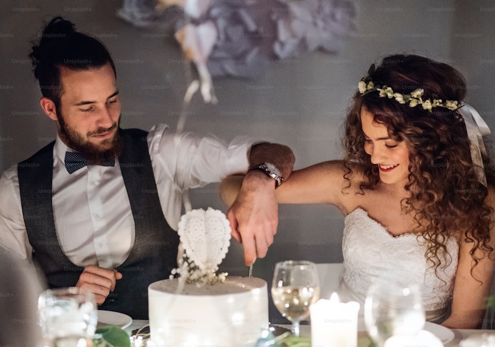 Um jovem casal feliz sentado em uma mesa em um casamento, cortando um bolo.