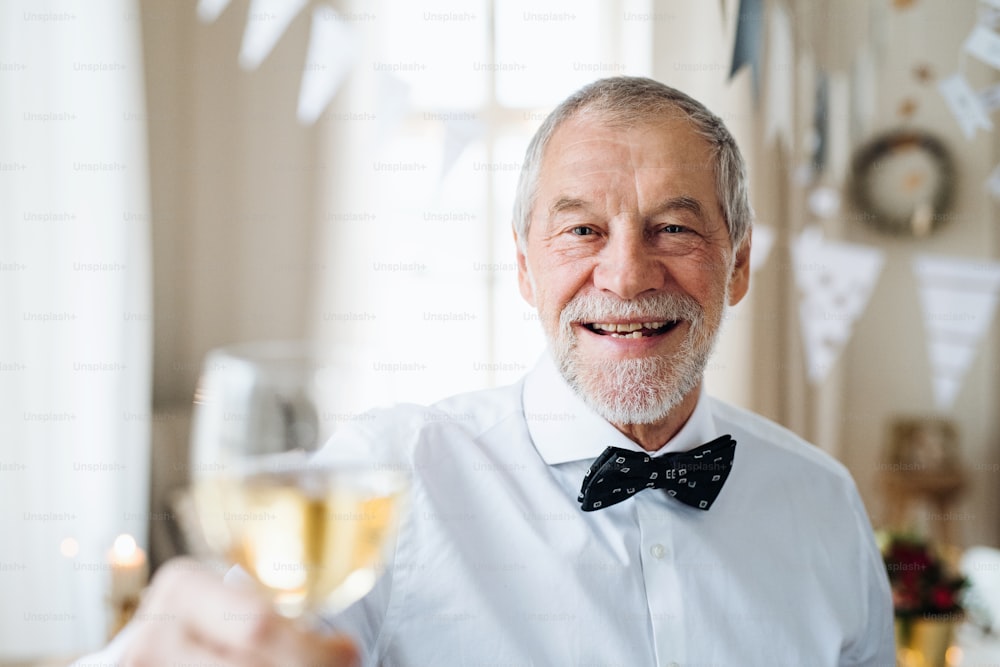 A portrait of a senior man standing indoors in a room set for a party, holding a glass of wine. Copy space.