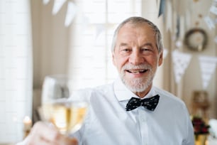 A portrait of a senior man standing indoors in a room set for a party, holding a glass of wine. Copy space.
