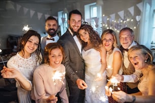젊은 신부, 신랑 및 다른 손님들이 결혼식 피로연에서 사진을 찍기 위해 포즈를 취하고 반짝임을 들고 있습니다.