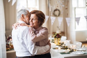 Portrait d’un couple de personnes âgées debout à l’intérieur dans une pièce aménagée pour une fête, s’étreignant.
