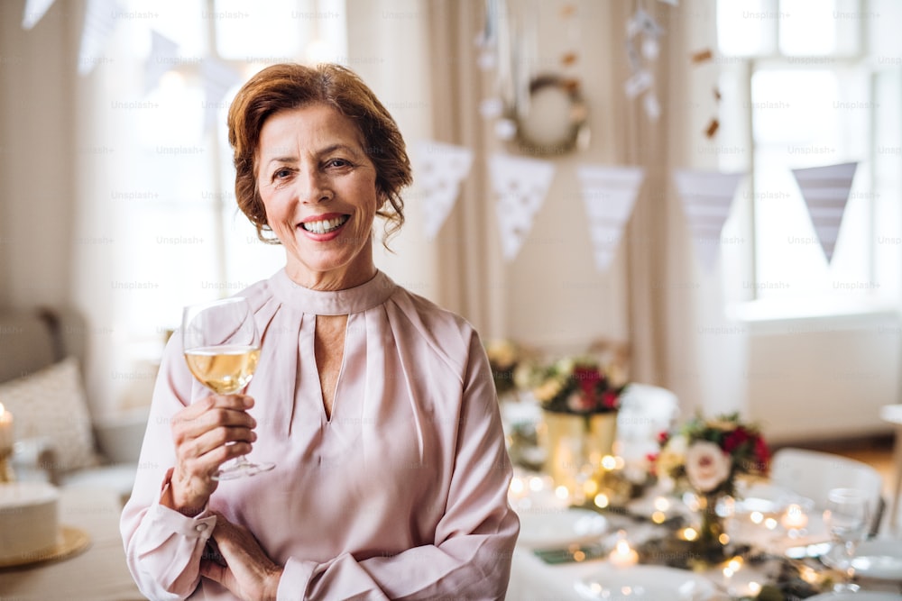 A portrait of a senior woman standing indoors in a room set for a party, holding a glass of wine. Copy space.