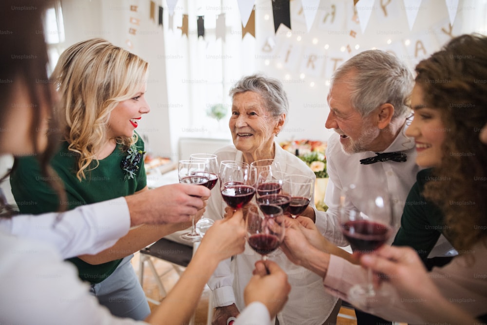 Una familia multigeneracional tintineando copas con vino tinto en una fiesta de cumpleaños familiar en el interior, haciendo un brindis.