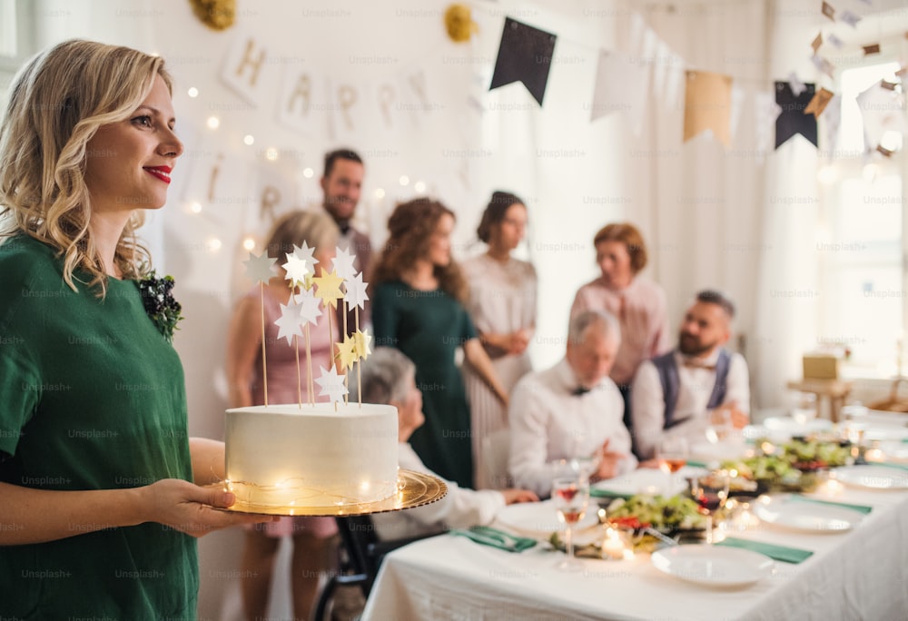 A young woman holding a birthday cake on an indoor party, multigeneration family in the background.