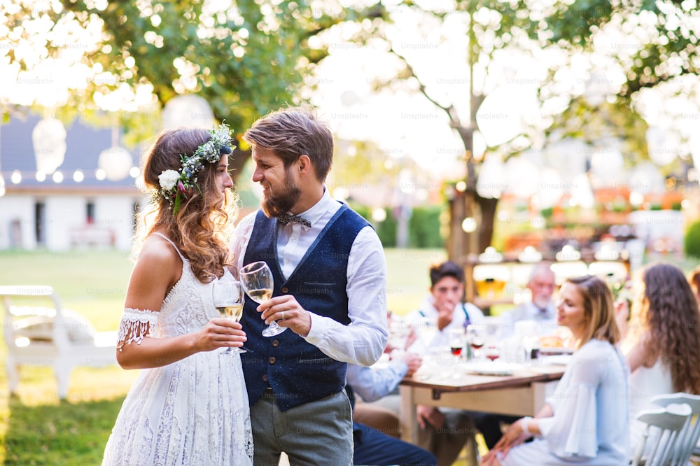 Wedding reception outside in the backyard. Bride and groom clinking glasses with champagne, guests sitting at the table in the background.