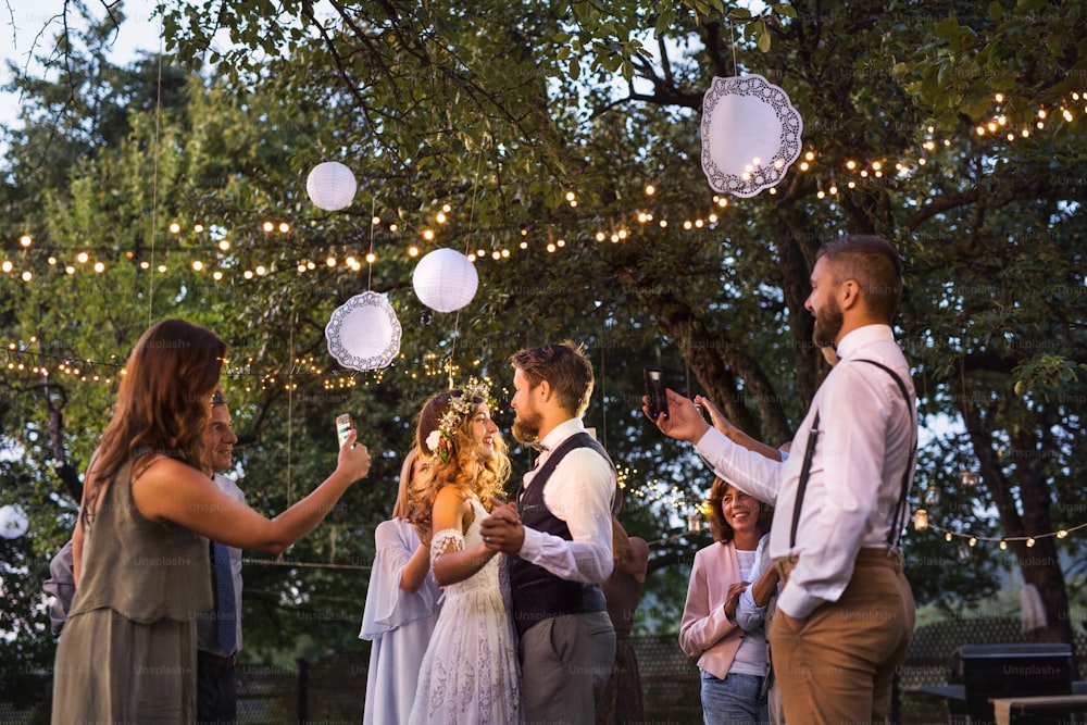 Gäste mit Smartphones, die ein Foto von einer tanzenden Braut und einem Bräutigam bei der Hochzeitsfeier draußen machen. Ein Abend im Hinterhof.