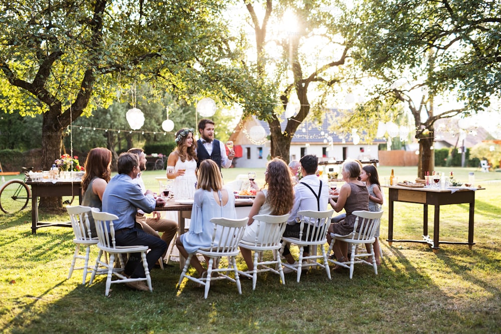 Recepción de boda afuera en el patio trasero. Guapo novio dando discurso. Invitados sentados alrededor de la mesa.