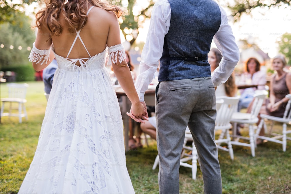 La mariée méconnaissable, le marié et les invités à la réception de mariage à l’extérieur dans la cour, se tenant la main. Vue arrière.