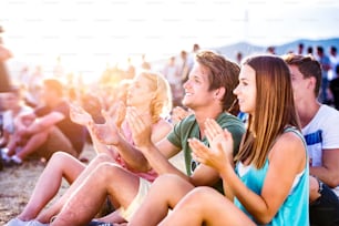 Gruppe von Teenagern auf dem Sommermusikfestival, auf dem Boden sitzend, Hipster-Junge im grünen T-Shirt
