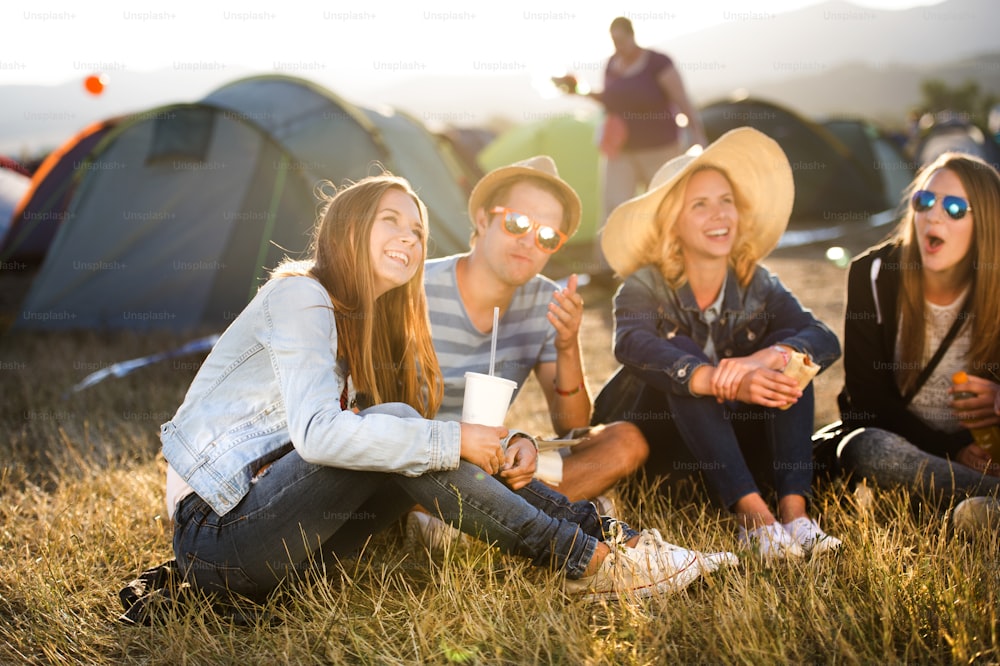 Group of teenage boys and girls at summer music festival, sitting on the ground in front of tents eating