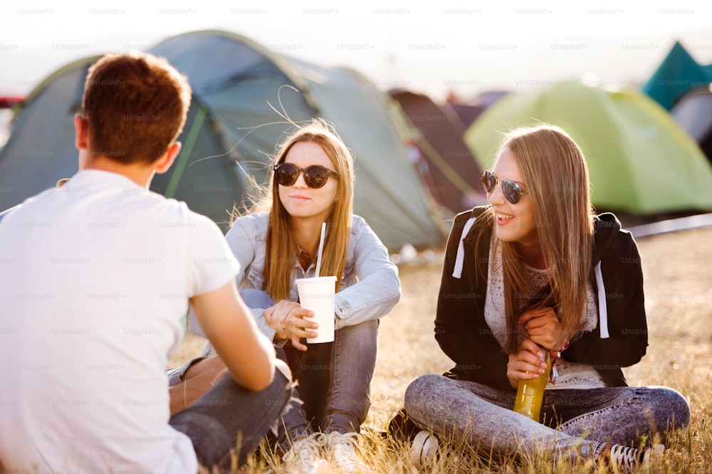 Group of teenage boys and girls at summer music festival, sitting on the ground in front of tents eating and drinking