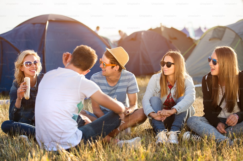 Group of teenage boys and girls at summer music festival, sitting on the ground in front of tents resting and eating