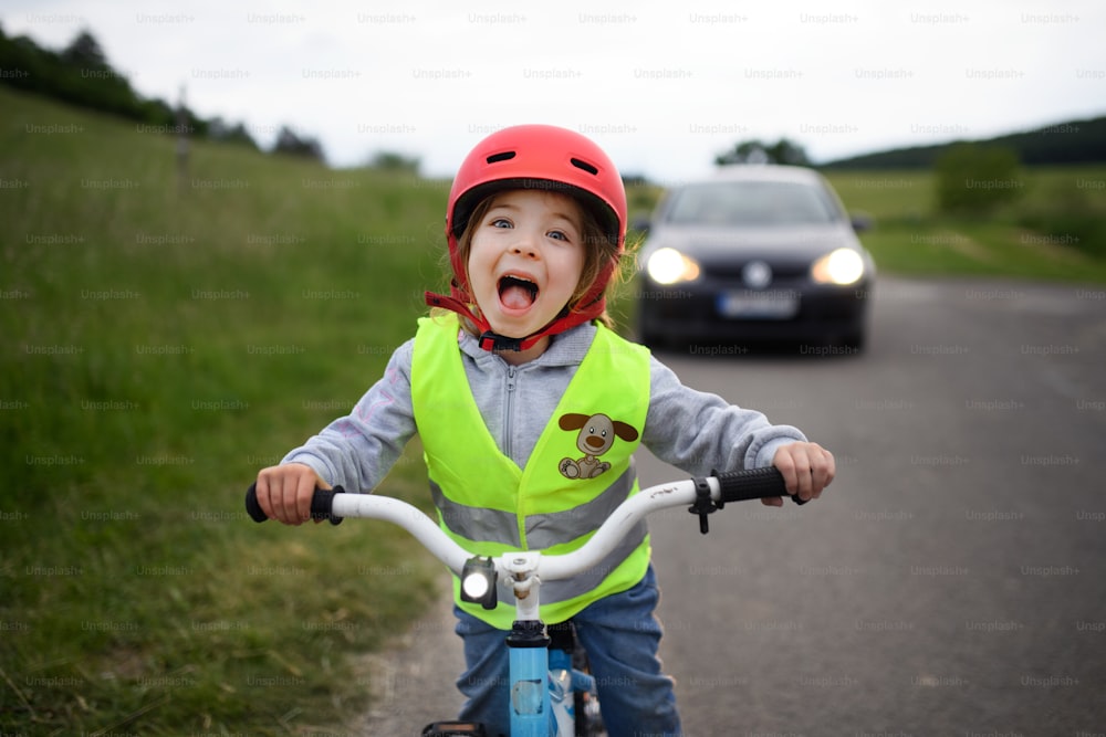 A portrait of excited little girl in reflective vest riding bike on road with car behind her