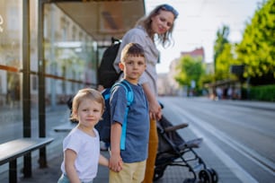 A young mother commuter with little kids on the way to school, waiting for bus in city.