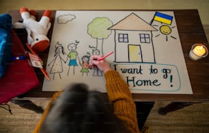 Eine Draufsicht auf ein ukrainisches Flüchtlingsschulmädchen, das sein Zuhause vermisst und ihre Familie zeichnet. Ukrainisches Kriegskonzept.