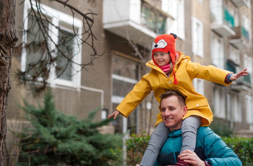 Ein Vater bringt seine kleine Tochter mit Down-Syndrom huckepack zur Schule, draußen auf der Straße.