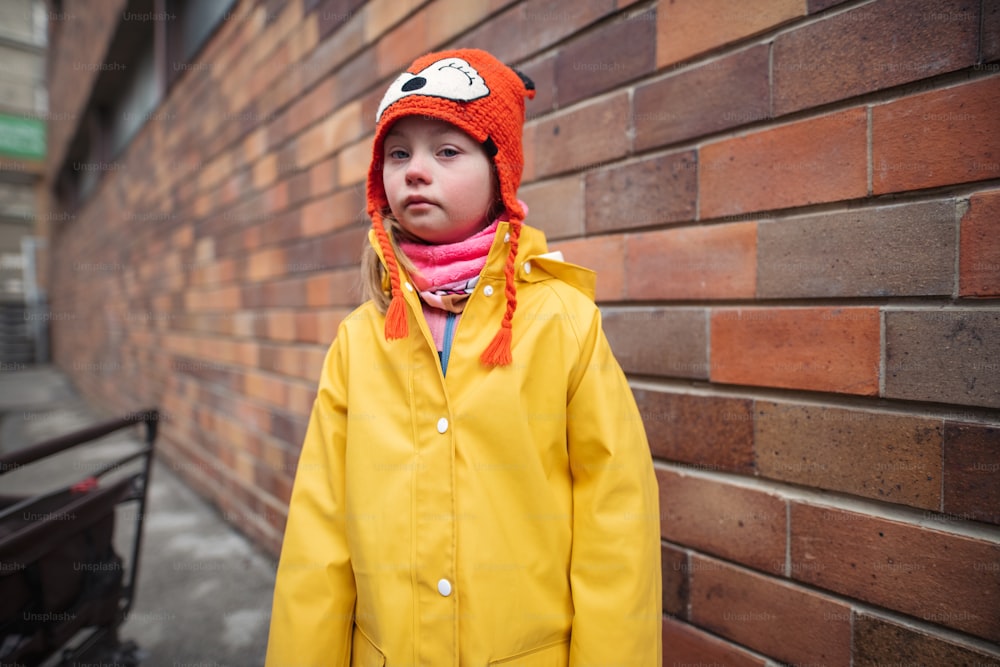 Uma garotinha com síndrome de Down olhando para a câmera outoors no inverno contra a parede de tijolos.