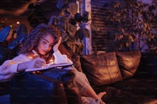 Eine glückliche junge Frau, die auf dem Sofa sitzt und abends im gemütlichen Wohnzimmer Tagebuch schreibt.