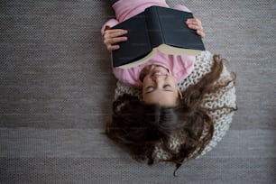 Eine Draufsicht auf ein glückliches kleines Mädchen, das auf dem Boden liegt und zu Hause ein Buch liest.
