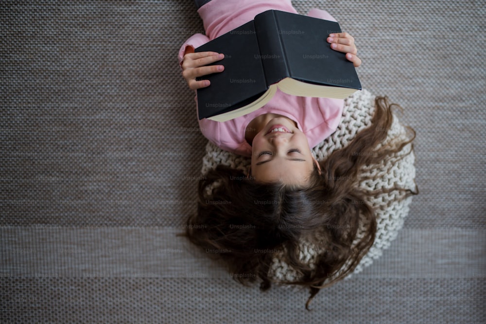 Una vista superior de una niña feliz acostada en el suelo y leyendo un libro en el interior de su casa.
