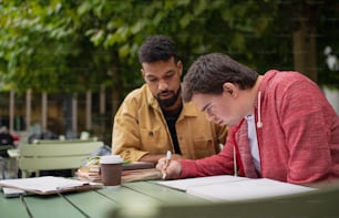 Un joven con síndrome de Down con su amigo mentor sentado al aire libre en un café y estudiando.