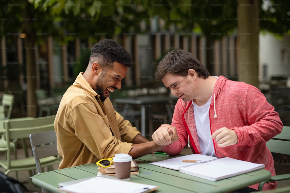 A young man with Down syndrome with his mentoring friend arm wrestling outdoors in cafe