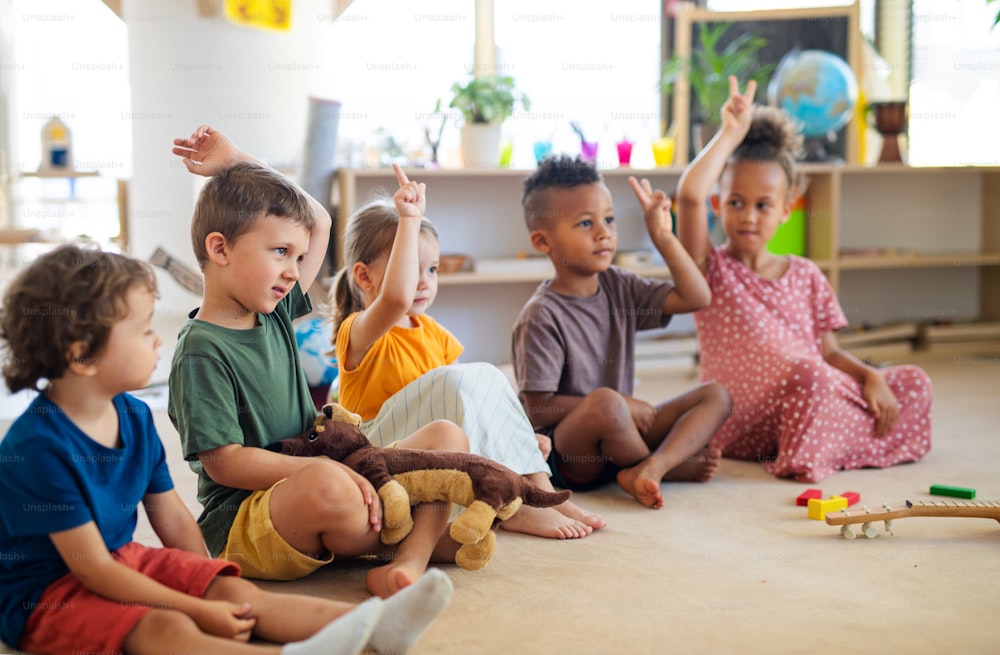 Um grupo de pequenas crianças da creche sentadas no chão dentro de casa de aula, levantando as mãos.