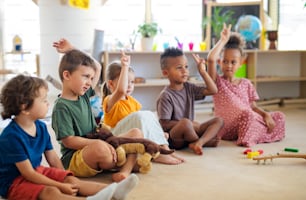 A group of small nursery school children sitting on floor indoors in classroom, raising hands.