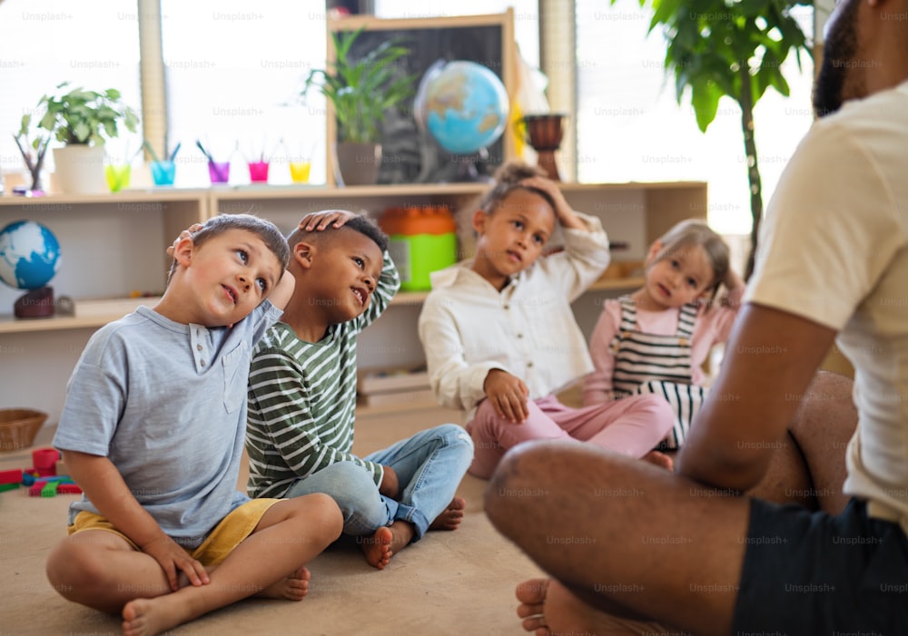 小さな保育園の子供たちのグループと、教室の屋内の床に座って運動をしている男性教師。