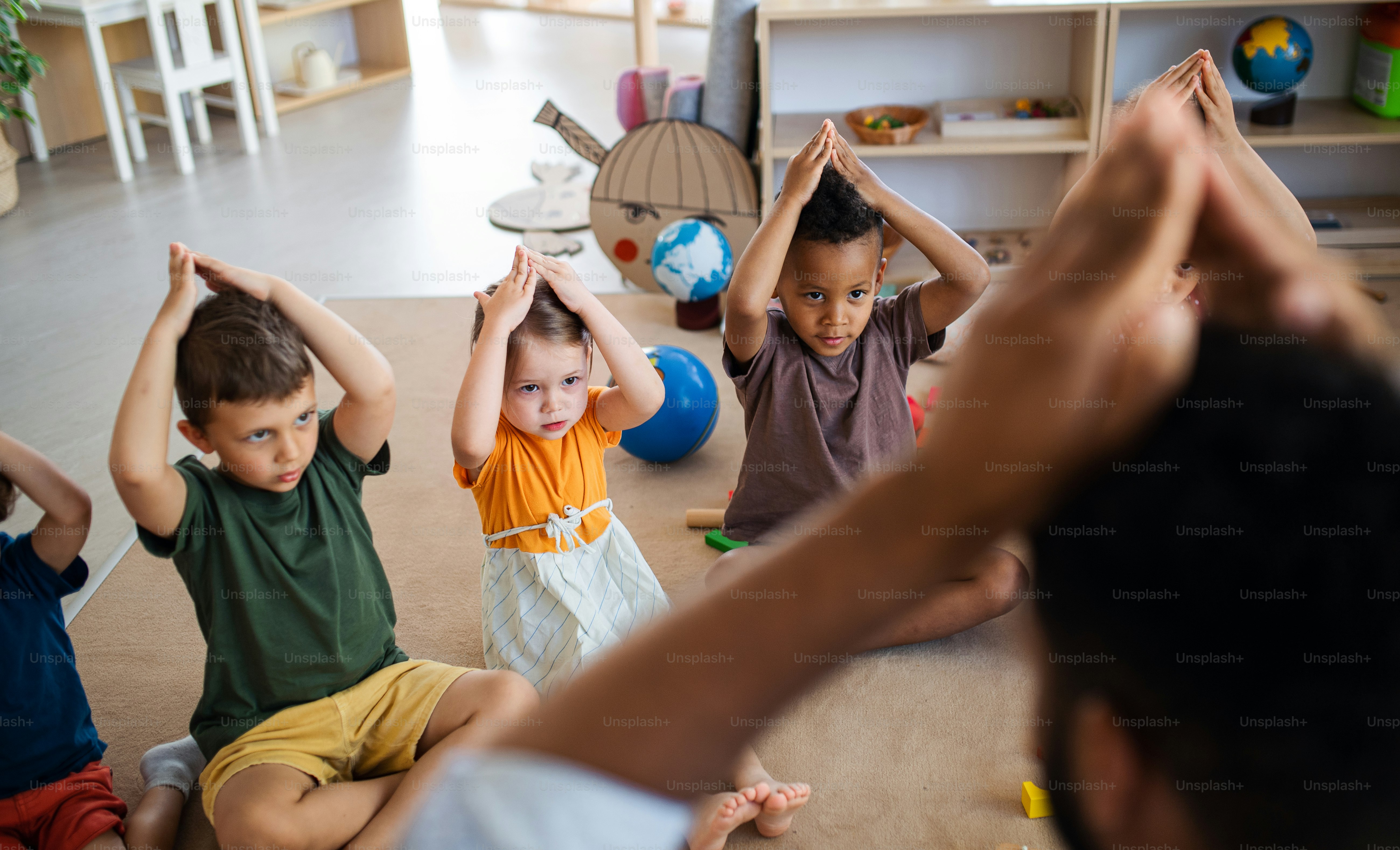 Eine Gruppe kleiner Kindergartenkinder mit einem Lehrer, der drinnen im Klassenzimmer auf dem Boden sitzt und spielt.