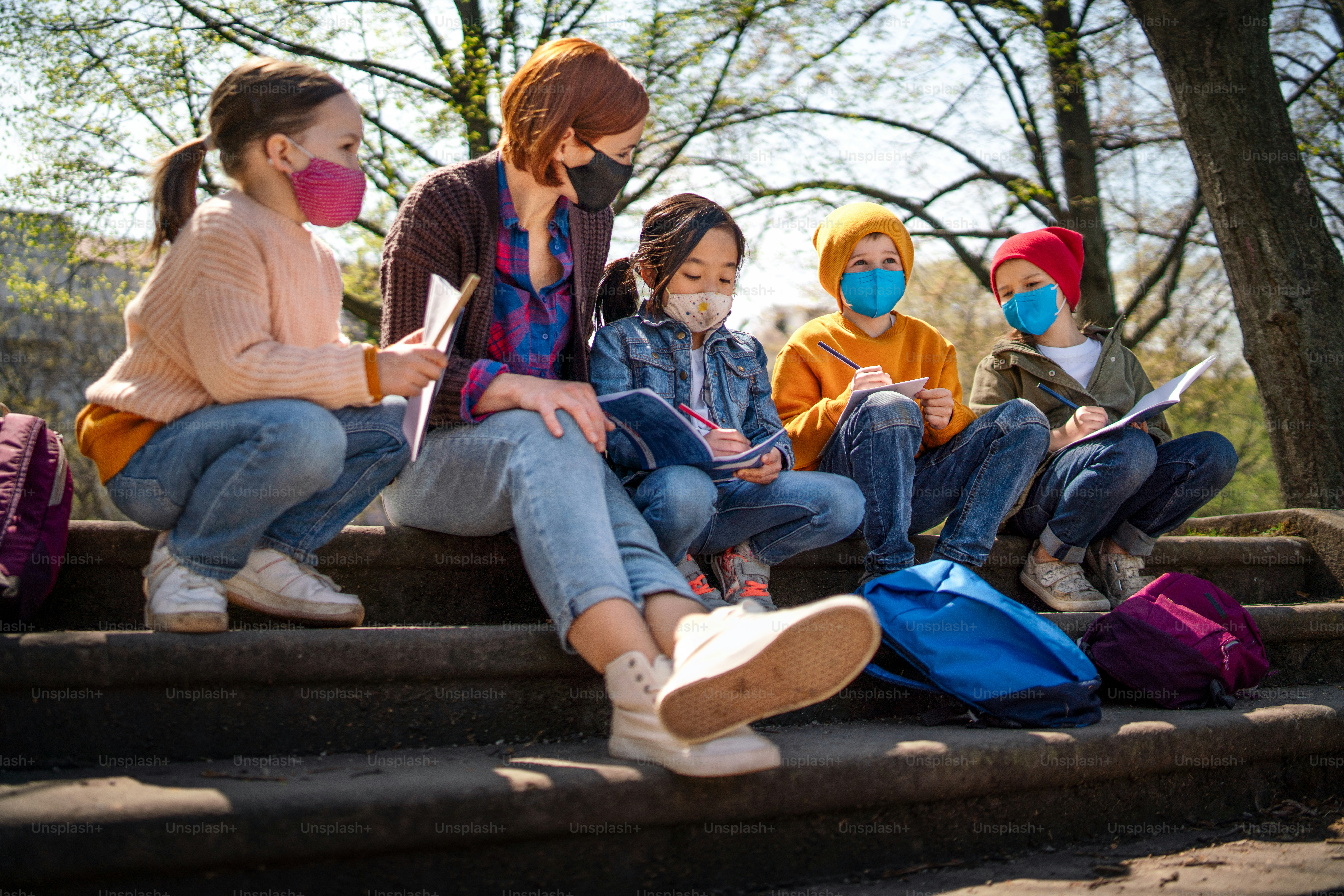 A teacher with small children sitting outdoors in city park, learning group education and coronavirus concept.