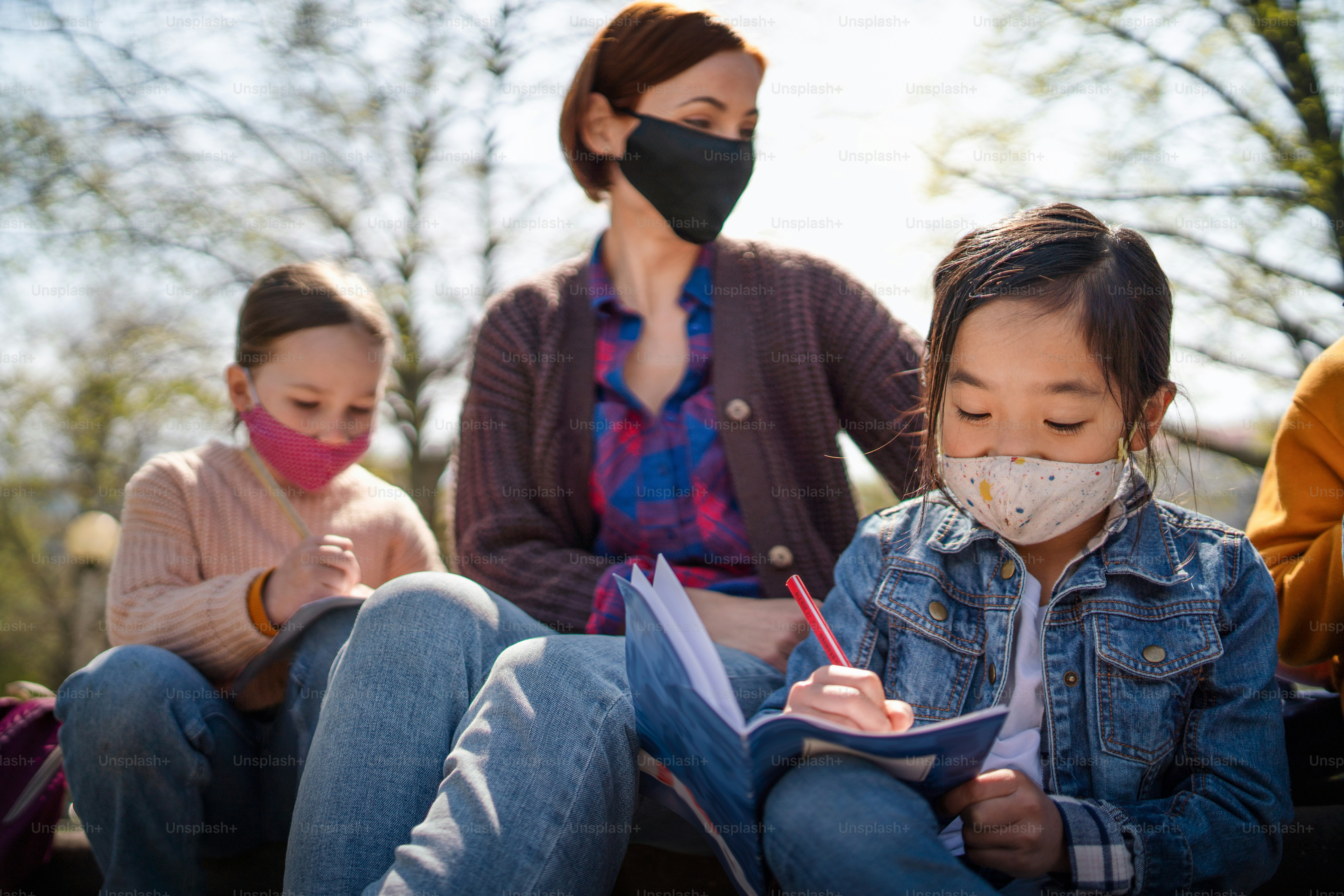 Uma professora com crianças pequenas sentadas ao ar livre no parque da cidade, aprendendo educação em grupo e conceito de coronavírus