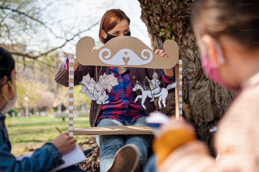 A teacher with small children sitting outdoors in city park, learning group education and coronavirus concept.