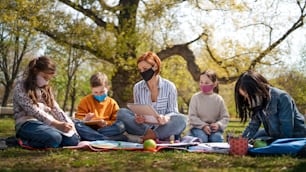 Uma professora com crianças pequenas sentadas ao ar livre no parque da cidade, aprendendo educação em grupo e conceito de coronavírus.