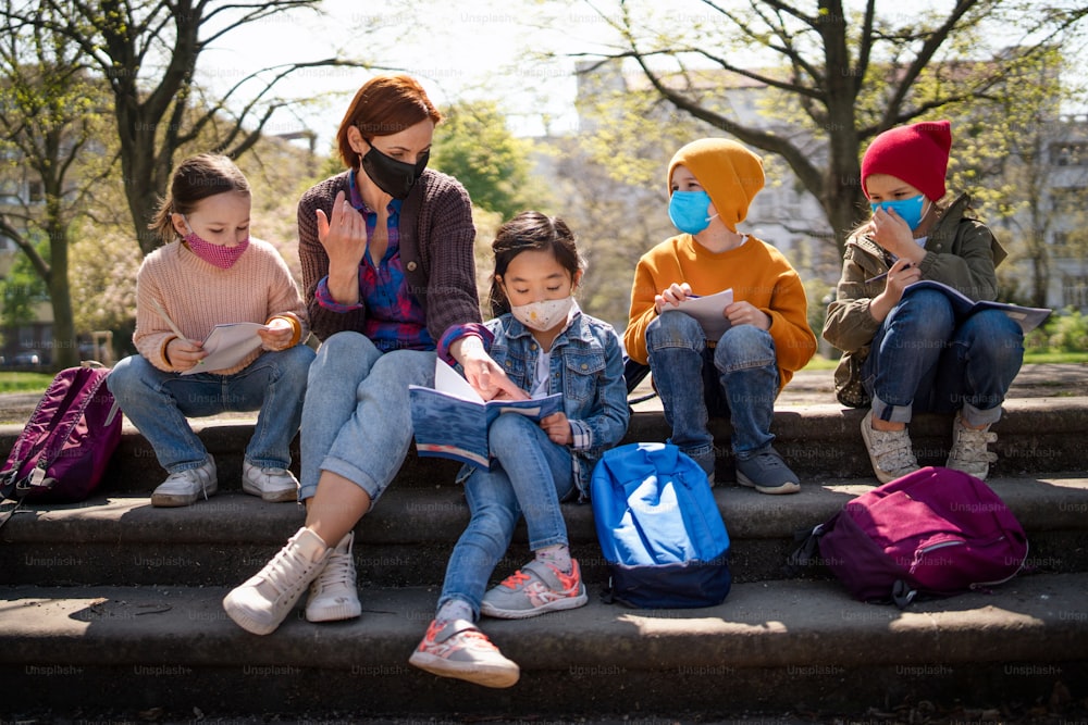 Un maestro con niños pequeños sentados al aire libre en el parque de la ciudad, aprendiendo educación grupal y concepto de coronavirus.