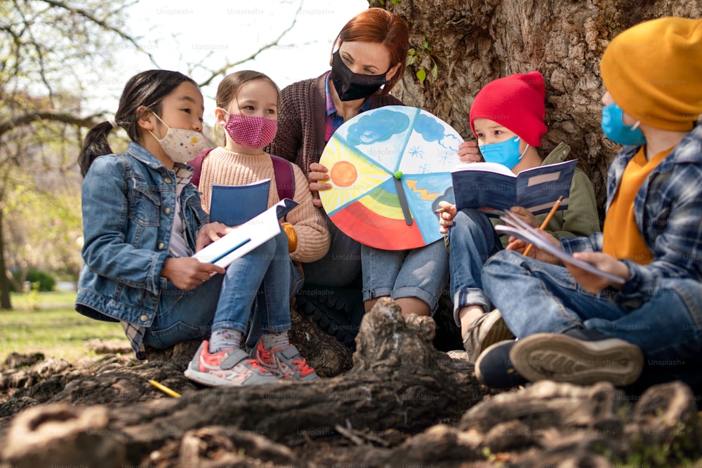 A teacher with small children sitting outdoors in city park, learning group education and coronavirus concept.