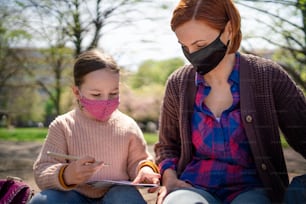 A teacher with small child sitting outdoors in city park, learning group education and coronavirus concept.