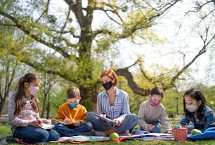 A teacher with small children sitting outdoors in city park, learning group education and coronavirus concept.