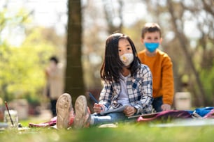 Crianças pequenas sentadas ao ar livre no parque da cidade, aprendendo educação em grupo e um conceito de coronavírus.