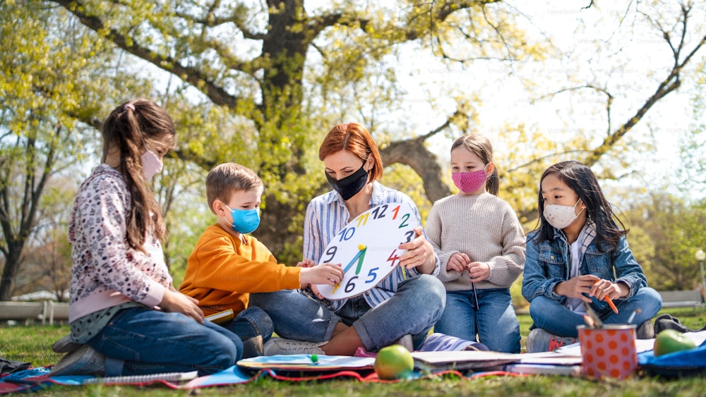 A teacher with small children sitting outdoors in city park, learning group education and coronavirus concept.