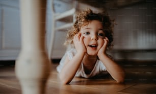 A portrait of cute small girl lying on floor under table indoors at home, looking away.