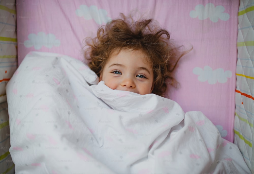 A top view of happy little girl lying in bed hiding under blanket, looking at camera indoors at home