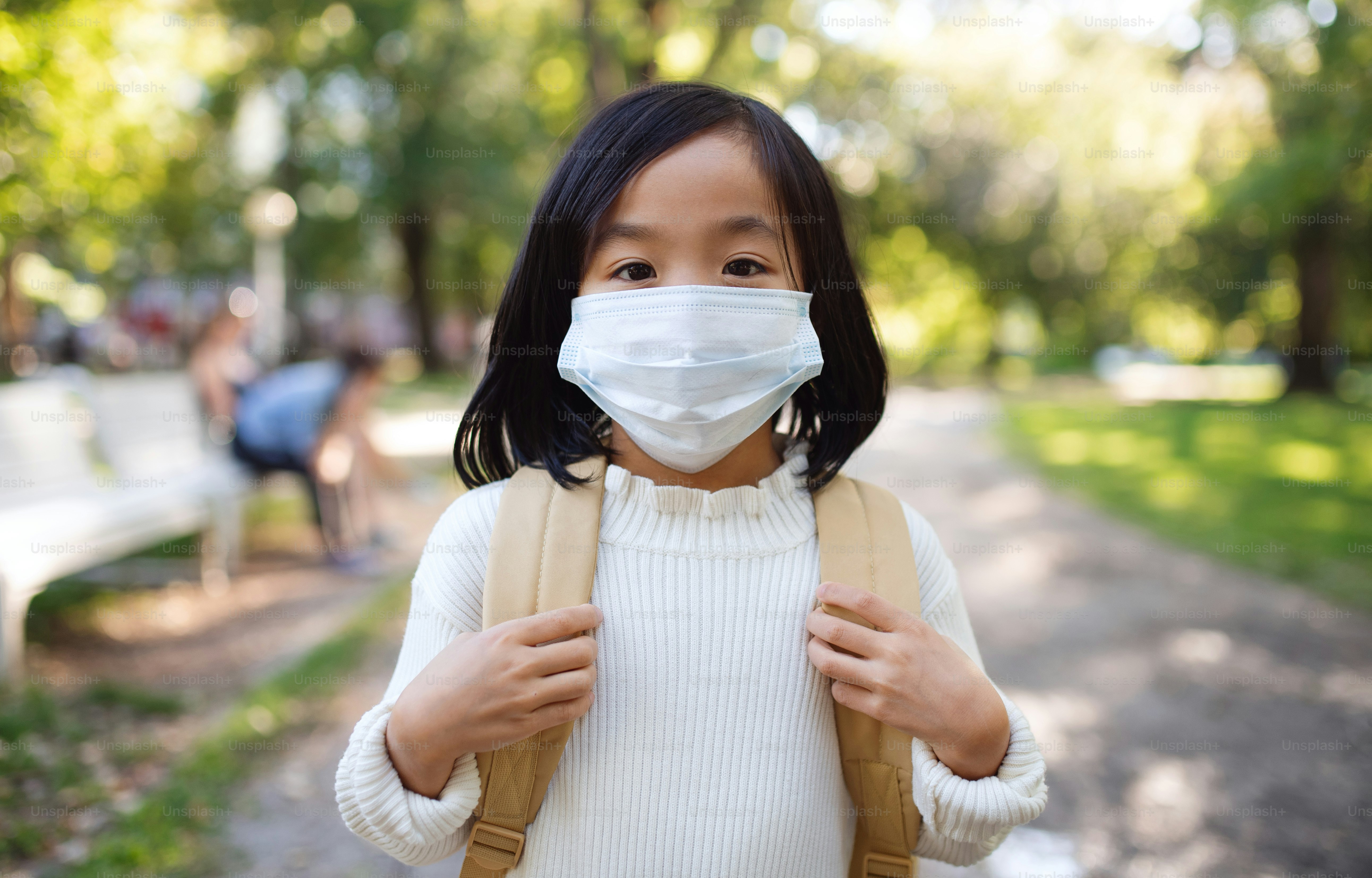 Front view portrait of small Japanese girl with backpack standing outdoors in town, coronavirus concept