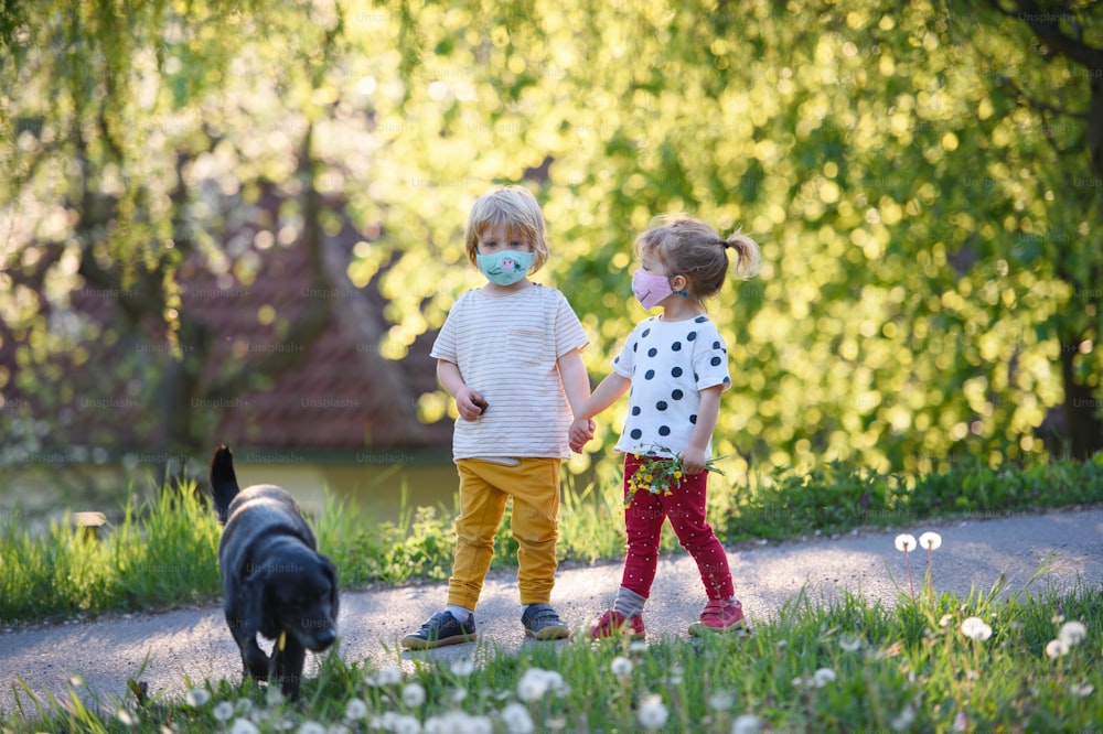 Petits enfants avec des masques faciaux et un chien jouant à l’extérieur à la campagne, concept de coronavirus.